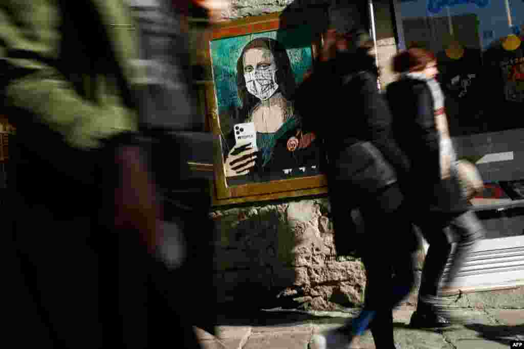 People walk by a poster by Italian urban artist Salvatore Benintende aka "TVBOY" depicting Leonardo da Vinci's Mona Lisa wearing a protective face mask and holding a mobile phone reading "Mobile World Virus" in a street of Barcelona, Spain.