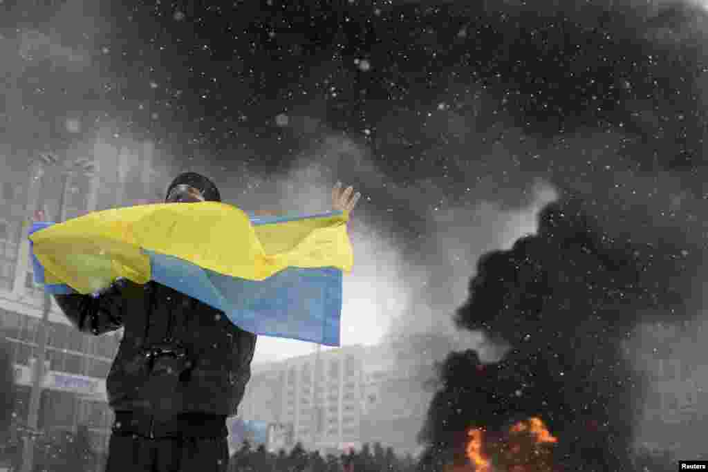 A man holds a Ukrainian flag as smoke rises in the background during clashes between police and pro-European protesters in Kyiv, Jan. 22, 2014.
