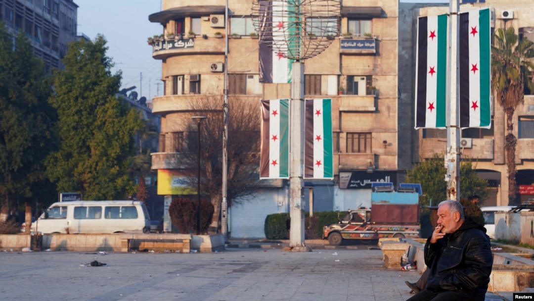 A man sits near flags placed by Syria's new leaders after the ousting of its longtime authoritarian President Bashar al-Assad, in Aleppo, Syria, Jan. 8, 2025.