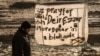 FILE - A man walks past a banner reading "Pray for Deir ez-Zor" at a migrant camp in Calais, France, Dec. 7, 2015. Years of conflict between Syrian government forces and Syrian opposition groups, and the emergence of extremist groups like Islamic State, have forced hundreds of thousands of people to flee from Deir ez-Zor province. 